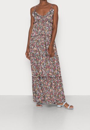 Femme portant une robe maxi sans manches à imprimé floral avec des détails à volants et des sandales à talons blocs ouvertes bleu clair sur un fond uni.