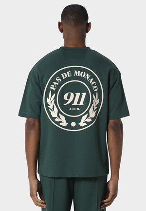 Homme portant un t-shirt vert foncé oversized avec un grand logo circulaire blanc au dos indiquant "Pas De Monaco 911 Club" entouré de feuilles de laurier.