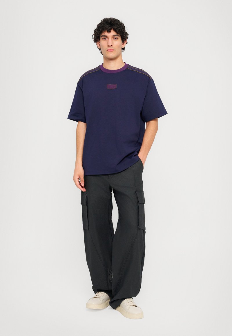 Navy t-shirt with purple collar, featuring a patch at the chest. Paired with loose-fitting black cargo pants and beige sneakers.
