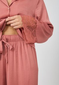 Ružový set na voľný čas s saténovým povrchom, ktorý sa skladá z blúzky s gombíkmi a širokými rukávmi. Ozdobený jemnou čipkou na manžetách a s nastaviteľným pásom na šnúrku.