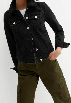 Zwarte spijkerjas met shearlingkraag, gedragen met een wit shirt en olijfgroene cargobroek, hand in de zak, tegen een witte achtergrond.