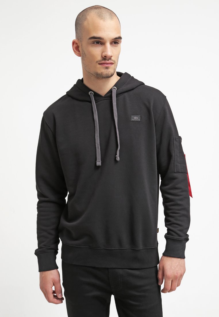 Sudadera negra con capucha ajustable, presenta un parche con el logo en el pecho y un acento rojo en el brazo izquierdo. Hecha de una tela suave y texturizada.