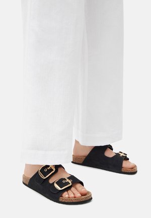 Schwarze Wildleder-Sandalen mit zwei breiten Riemen und goldenen Schnallen. Kork-Fußbett unter der Sohle für Komfort; kombiniert mit weißen Stoffhosen.