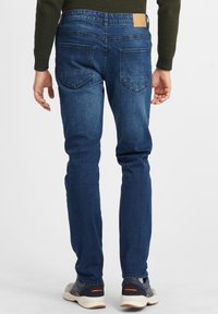Homme portant un jean slim-fit bleu foncé, un pull vert olive et des baskets noir et blanc, se tenant de dos face à l'appareil photo sur un fond blanc.