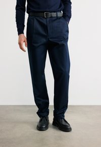 Άνδρας που φοράει μαύρη δερμάτινη ζώνη, ναυτικό μπλε πλισέ παντελόνια, μαύρα δερμάτινα παπούτσια, στέκεται με το ένα χέρι στην τσέπη σε απλό φόντο.