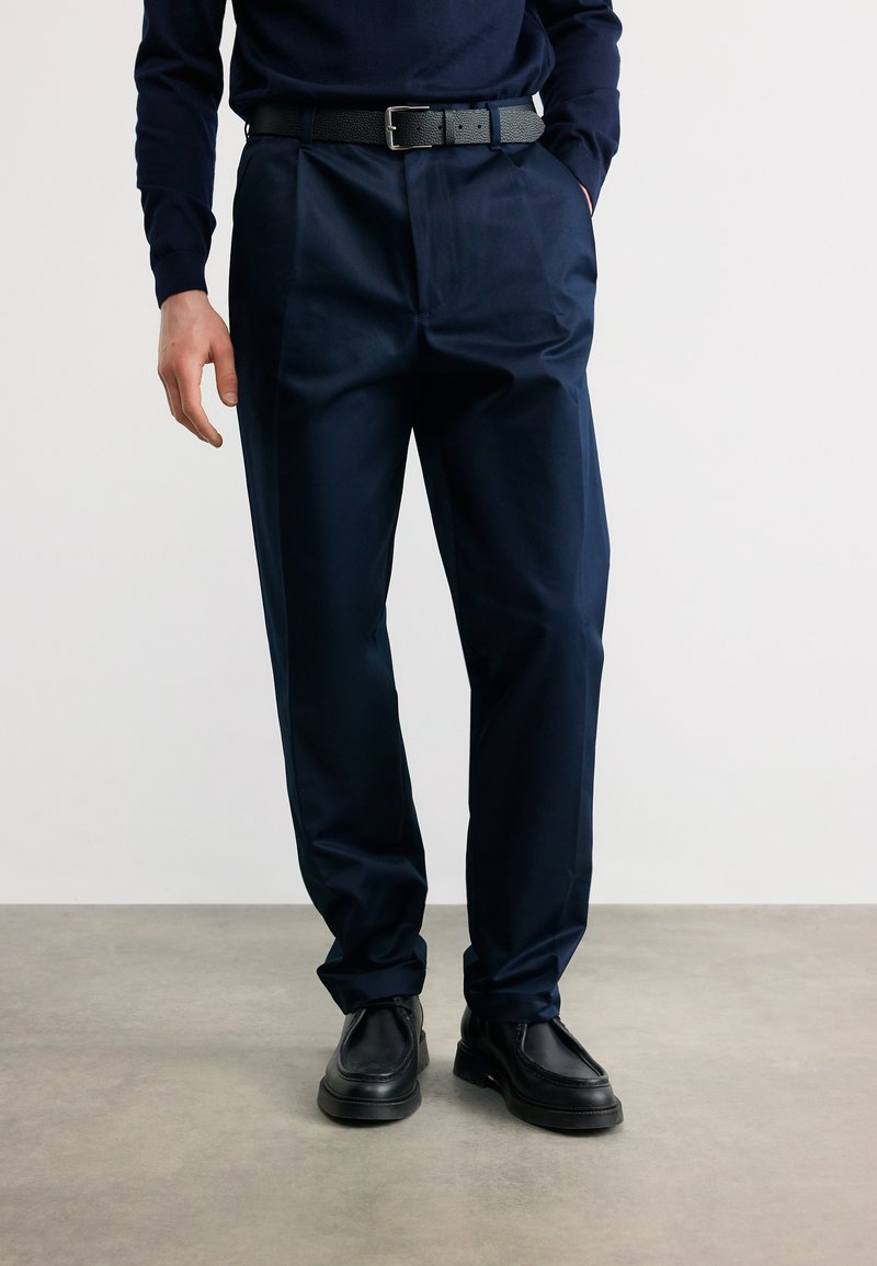 Άνδρας που φοράει μαύρη δερμάτινη ζώνη, ναυτικό μπλε πλισέ παντελόνια, μαύρα δερμάτινα παπούτσια, στέκεται με το ένα χέρι στην τσέπη σε απλό φόντο.