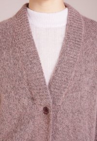 Ljusrosa kofta i mjukt, fluffigt material med ribbad krage, enstaka knappstängning och en lager vit tröja under.