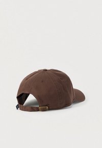 Καφέ βαμβακερό καπέλο με καμπυλωτό γείσο, ρυθμιζόμενο λουράκι στο πίσω μέρος και μικρή υφασμένη ετικέτα. Διαθέτει ομοιόμορφη υφή χωρίς ορατά σχέδια.