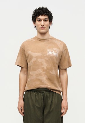 Junger Mann mit lockigem Haar trägt braunes T-Shirt mit Camouflage-Muster „Aries“ und dunkelgrüne Hose, stehend vor neutralem Hintergrund.