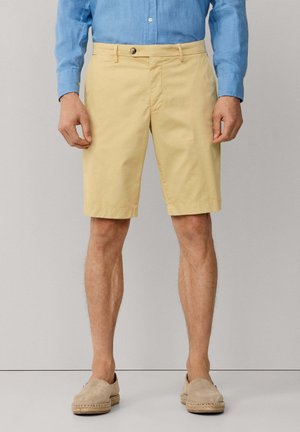 Homme portant un short jaune clair jusqu'aux genoux, une chemise à manches longues bleue et des chaussures beige à enfiler, debout devant un fond uni.