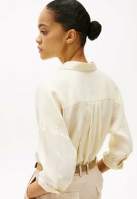 Crème-kleurig, getextureerd shirt met een kraag en geplooide achterkant. Ruime pasvorm, opgerolde mouwen, gecombineerd met beige broek.