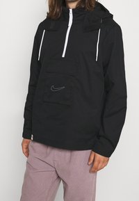 Black, lightweight anorak jacket with a half-zip, front pocket, and white accents. Features a hood and subtle Nike logo embroidery.