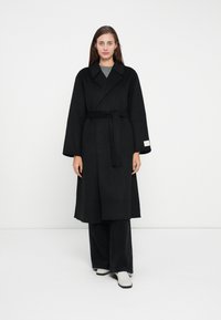Long black coat made of a thick fabric, featuring wide sleeves, a tie belt, and a collar. Worn over gray top with flared black pants.