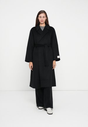 Long black coat made of a thick fabric, featuring wide sleeves, a tie belt, and a collar. Worn over gray top with flared black pants.