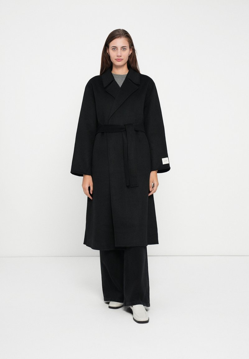 Long black coat made of a thick fabric, featuring wide sleeves, a tie belt, and a collar. Worn over gray top with flared black pants.