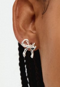 Silver bow-shaped earring with a polished finish, featuring flowing ribbons and a textured center, attached to a pierced ear.