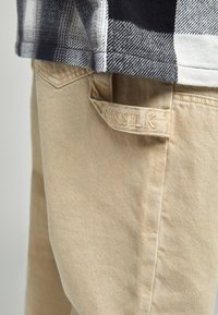 Pantaloni cargo beige con una finitura testurizzata, caratterizzati da un'etichetta in vita visibile con il marchio "SikSilk". Il tessuto appare resistente e casual.