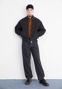 Svart bomberjakke med ribbede mansjetter, brun turtleneck og mørke vide bukser. Accessoriesert med en svart lue og kraftige svarte støvler.