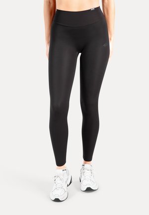Schwarze, hoch taillierte Leggings aus glattem, dehnbarem Stoff. Verfügen über ein eng anliegendes Design und ein Logodetail am Bund. Kombiniert mit weißen Turnschuhen.