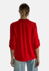 Camicia rossa a maniche lunghe con polsini arrotolati, caratterizzata da una leggera lucentezza e una piega sul retro. Abbinata a jeans blu chiaro.