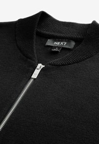 Pull noir à fermeture éclair avec col et poignets côtelés, muni d'une fermeture éclair argentée. L'étiquette indique "NEXT", fabriqué au Bangladesh. Texture tricotée douce.