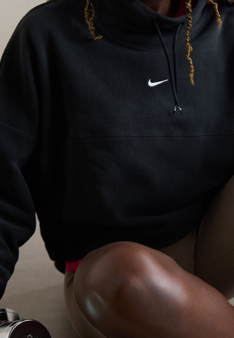 Personne portant un sweat à capuche Nike noir et un pantalon beige, assise avec un genou relevé, tenant un haltère argenté près du sol.
