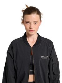 Junge Frau mit heller Haut und zurückgebundenem Haar, trägt eine schwarze Reißverschlussjacke über einem schwarzen Crop-Top mit "HALO"-Logo.