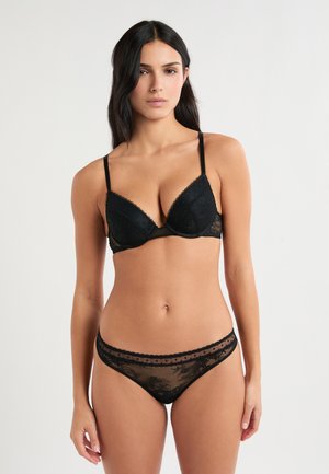 Mujer con cabello largo y oscuro, vestida con un sujetador negro de encaje con tirantes finos y bragas a juego de encaje, de pie contra un fondo blanco liso.