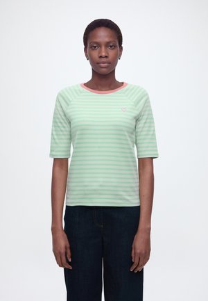 Femme aux cheveux courts portant une chemise rayée vert menthe et blanc avec un col rose et un pantalon bleu foncé, debout devant un fond uni.