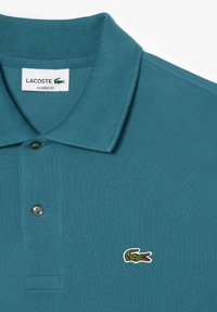 Teal-paitainen polo, joka on valmistettu teksturoidusta kankaasta ja jossa on ribbiriippuvainen kaulus, kaksinappinen nappilista ja pieni brodeerattu krokotiililogo.