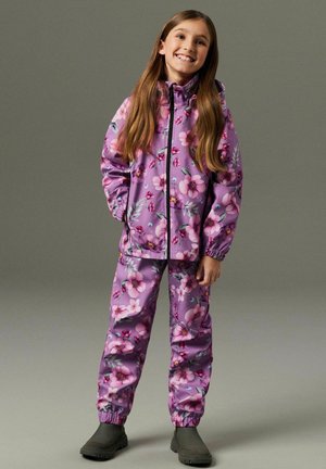 Jente som smiler med langt hår, iført en lilla jakke med blomsterprint og matchende bukser, stående med hendene i lommene og grå støvler.