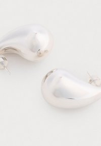 Srebrne, metaliczne kolczyki w gładkim, zakrzywionym kształcie kropli i z zapięciem na wkręty. Błyszczące wykończenie bez widocznych wzorów czy akcentów.