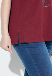 Camisa de manga corta burdeos con aberturas laterales, combinada con jeans de mezclilla azul. La tela parece suave y tiene una textura blanda.