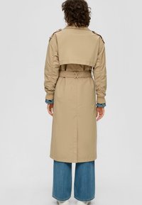 Beżowy trenchcoat z paskiem w talii, skróconym przodem, detalami na rękawach i rozporkiem z tyłu, noszony na szerokich dżinsowych spodniach.