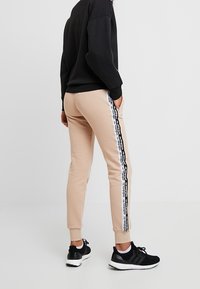 Beige träningsoveraller med smal passform, prydda med svarta sidestreck och logodetaljer, kombinerade med svarta sportskor och en svart sweatshirt.