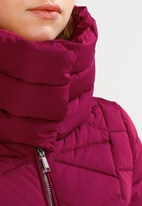 Primer plano de una persona vistiendo una chaqueta de plumas magenta con cuello alto y acolchado, con un cierre plateado visible, contra un fondo liso.