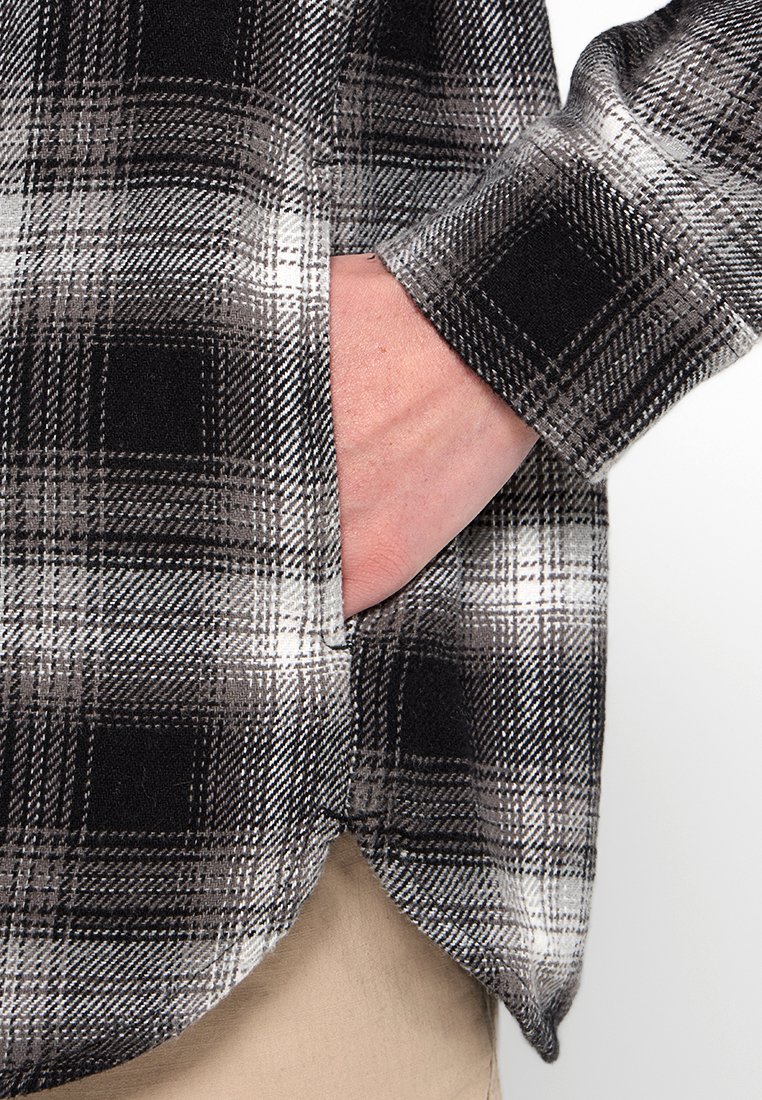 Eine Hand steckt in der Seitentasche eines schwarz-weißen karierten Hemdes, das über beigen Hosen getragen wird.