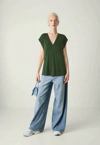 Top verde a V con maniche a cappuccio abbinato a jeans larghi blu chiaro. Modella che tiene una piccola borsa blu, sneakers bianche in vista.