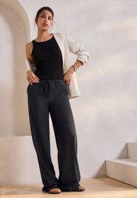 Canotta nera, blazer beige chiaro e pantaloni neri larghi con vita elastica, abbinati a sandali neri bassi. Design minimalista e rilassato.