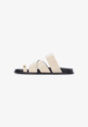 Sandalo piatto beige con suola nera, caratterizzato da tre cinturini, incluso un anello per l'alluce con dettaglio di bottone dorato.