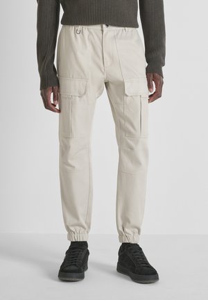 Pantalones cargo beiges con múltiples bolsillos, puños elásticos y cintura con cordón, combinados con zapatillas negras. Textura suave y ajuste entallado.