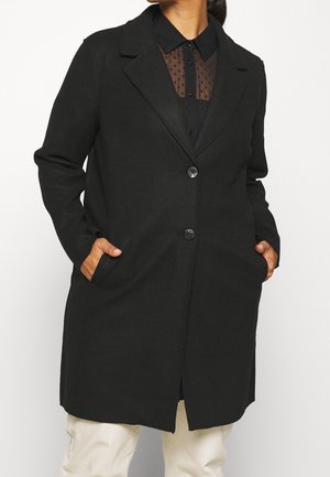 Femme portant un long manteau noir par-dessus un chemisier noir transparent à pois, les mains dans les poches du manteau et portant un pantalon blanc.