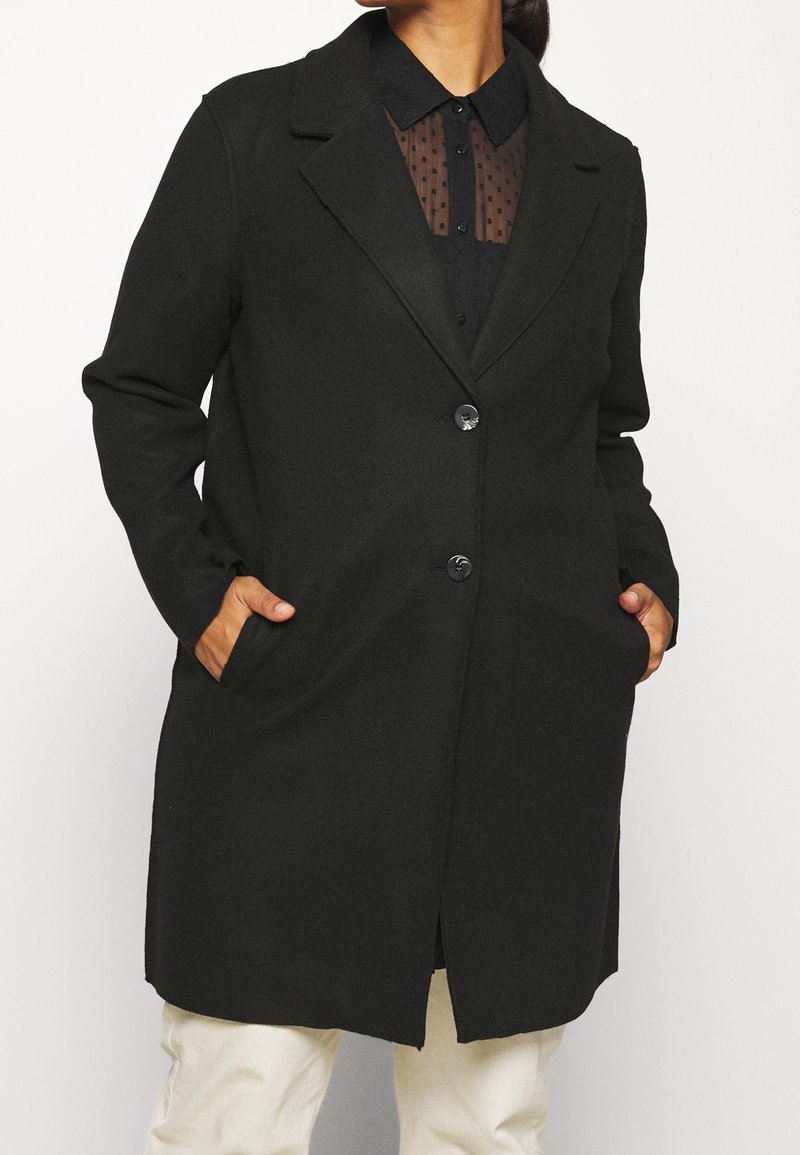 Donna con un lungo cappotto nero su una blusa nera trasparente con pois, con le mani nelle tasche del cappotto e pantaloni bianchi.