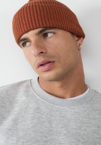 Rdzawy, prążkowany beanie, o miękkiej teksturze, noszony z jasnoszarym dresowym swetrem. Posiada zaokrąglony szczyt i widoczną obręcz na uszy.