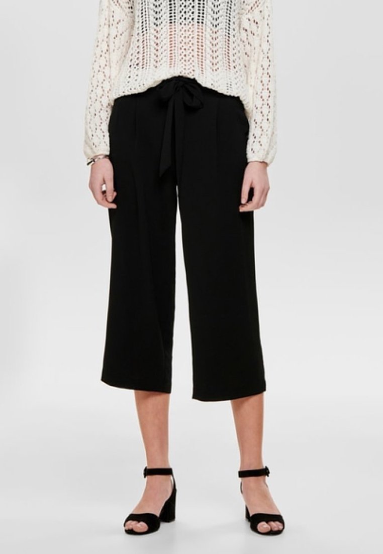 Culottes noires à jambes larges avec taille à nouer. Tissu lisse, longueur cheville. Associées à un haut blanc en maille à manches longues et des sandales noires à talons.