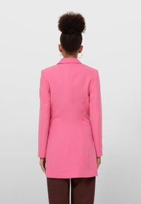 Rosa maßgeschneiderter Blazer mit langen Ärmeln, strukturierten Schultern, einer Naht auf der Rückseite und ohne sichtbare Verschlüsse. Getragen über braunen Hosen.