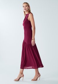 Bordo midi suknelė, pagaminta iš lygaus audinio, su prigludusiu liemeniu, liemens raukiniais ir platėjančia apačia. Skaidrios aukštakulnės sandalai.