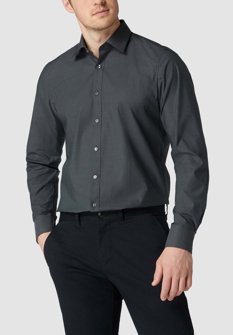 Homme portant une chemise boutonnée à manches longues gris foncé et un pantalon noir, les mains partiellement dans les poches, sur un fond clair uni.
