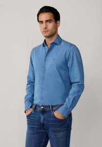 Lichtblauw overhemdd met lange mouwen, voorzien van een puntige kraag en subtiele logo-detail, gecombineerd met donkerblauwe jeans.