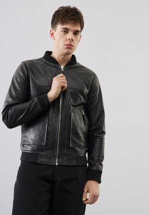 Junger Mann trägt eine schwarze Lederjacke mit Reißverschluss vorne und Taschen, hält den Reißverschluss an der Brust und steht vor einem grauen Hintergrund.